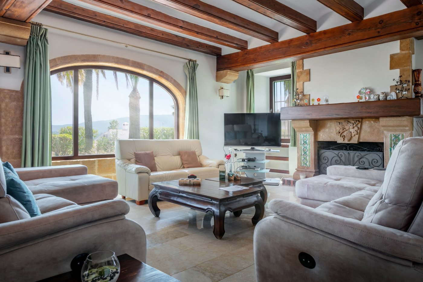 Woonkamer in mediterrane stijl met houten balkenplafond, groot boogvenster van Tosca-steen en een rustieke open haard met uitzicht op de vallei.