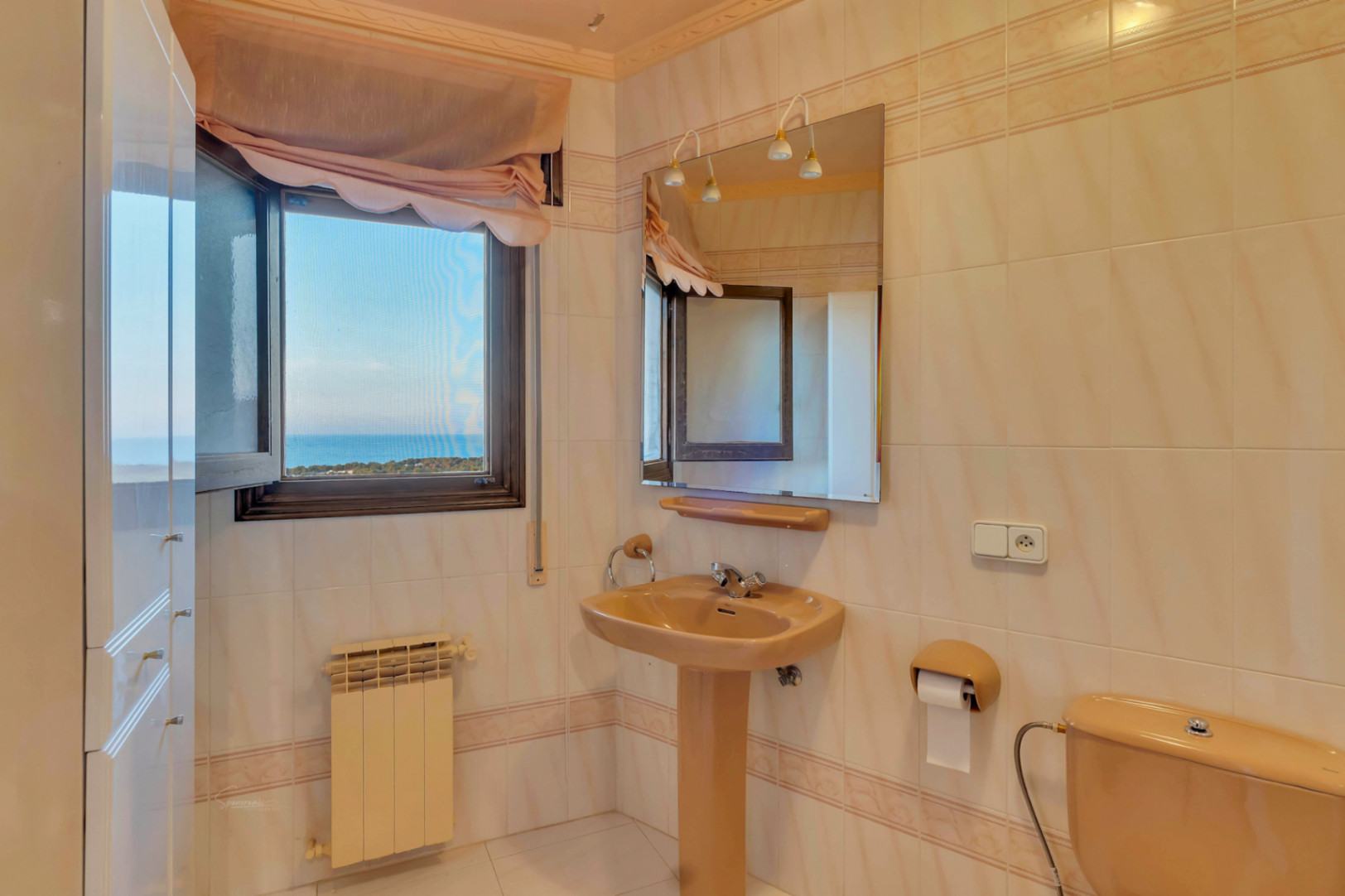 Badkamer met beige sanitair, een kolomwastafel en wandspiegel. Het venster biedt een helder uitzicht op de Middellandse Zee.