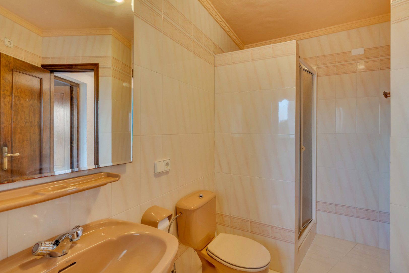 Badkamer met crème wandtegels, perzikkleurig sanitair en een inloopdouche met glazen deur. Voorzien van een grote spiegel en klassieke details.