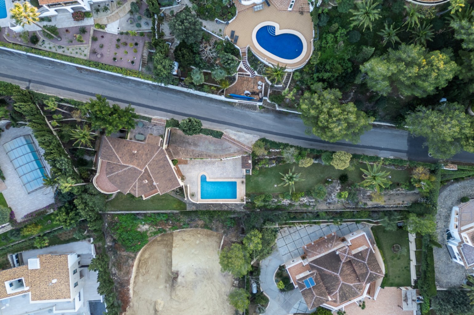 Bovenaanzicht van de villa met de indeling van het perceel, het rechthoekige zwembad, de geplaveide terrassen en de mediterrane tuin met palmbomen.