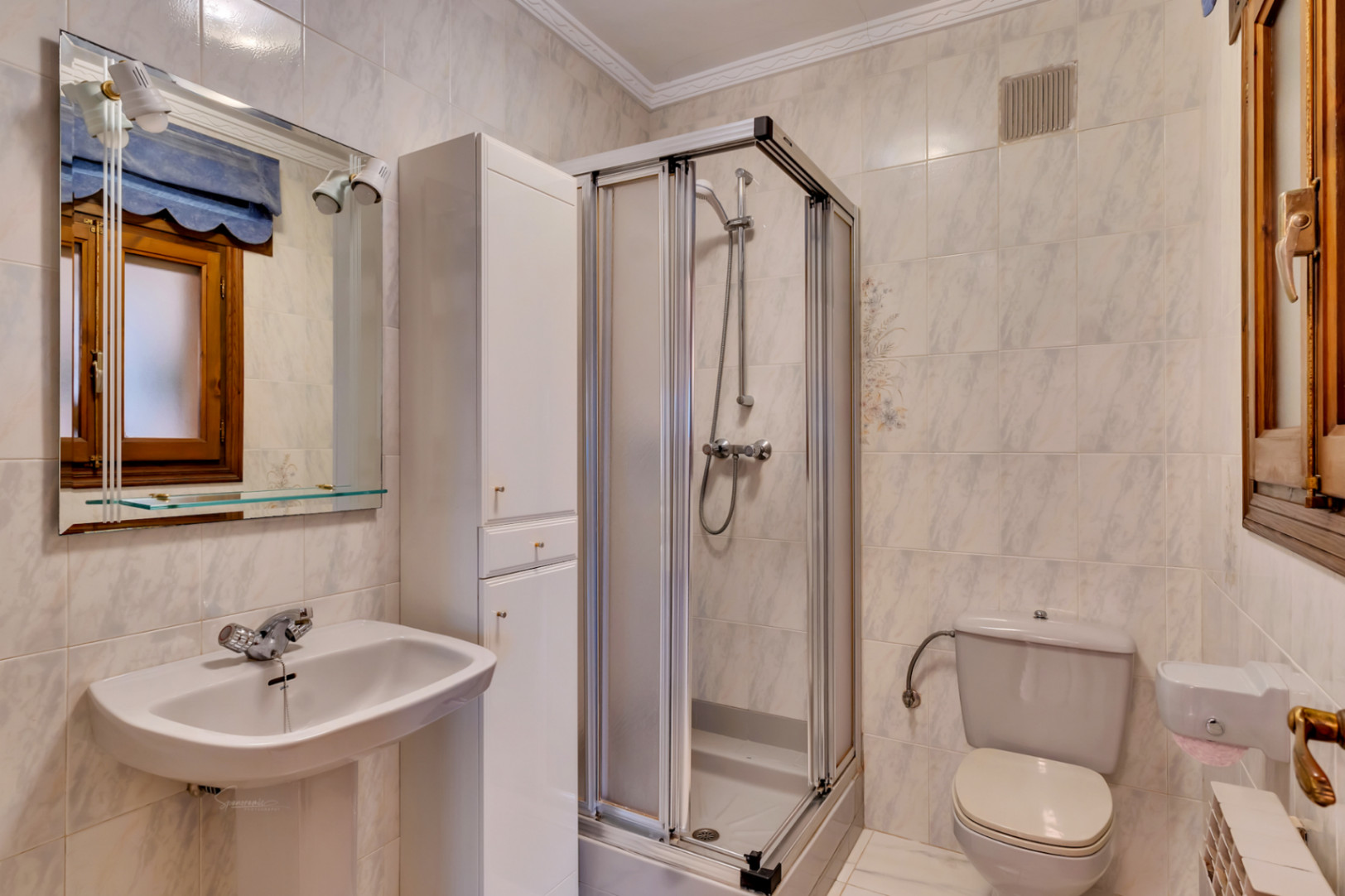 Functionele badkamer met een wastafel op zuil, een hoekdouchecabine met schuifdeuren en een hoge witte opbergkast. De wanden zijn bekleed met tegels in marmerlook.