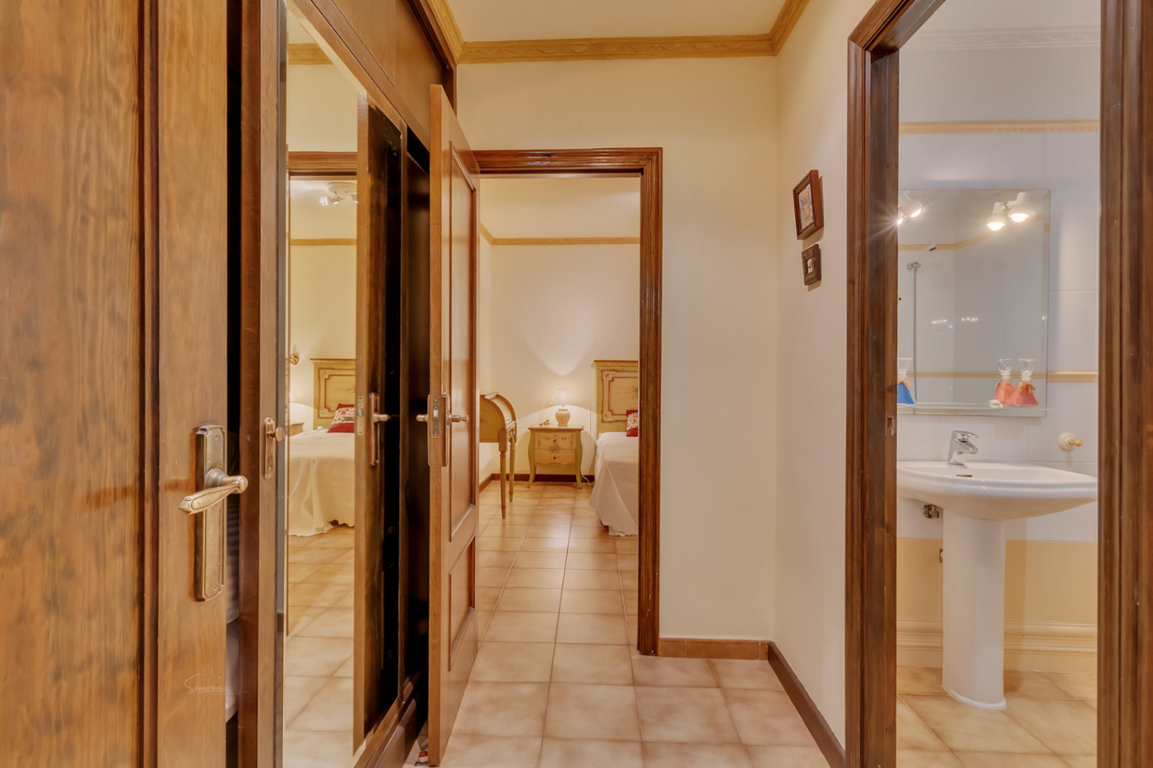 Gang met keramische tegelvloer die toegang biedt tot een slaapkamer met twee eenpersoonsbedden, een badkamer met kolomwastafel en houten inbouwkasten met spiegels.