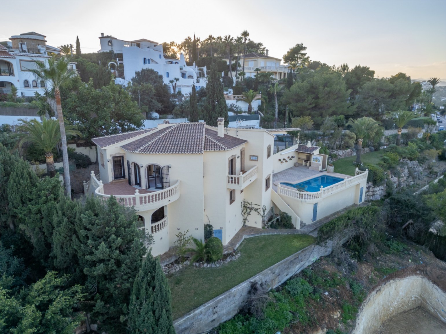 Luchtfoto van de villa met crèmegevel, terracotta dak en gebogen terrassen. De buitenruimte is voorzien van een zwembad, barbecueplaats en mediterrane tuin.