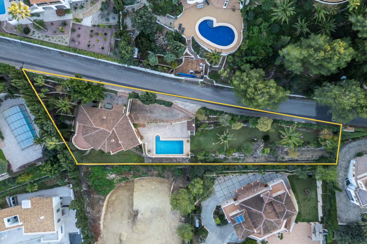 Luchtfoto van het omheinde perceel met het L-vormige zwembad, de mediterrane tuin met palmbomen en de villa met haar kenmerkende terracotta pannendaken.