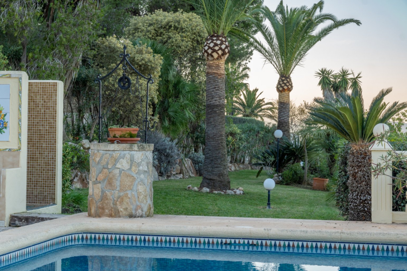 Mediterrane tuin met gazon, palmbomen en een decoratieve stenen put. Het zwembadgedeelte bevat een buitendouche met mozaïektegels en bolvormige tuinverlichting.