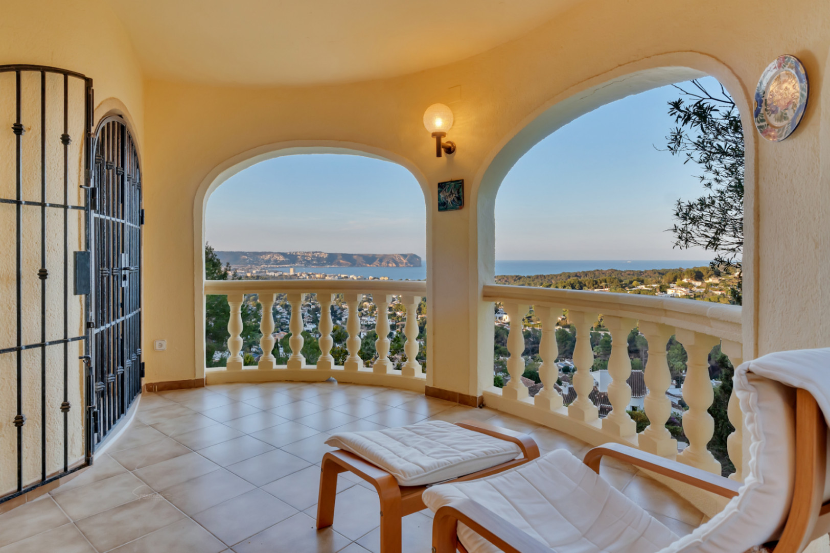 Overdekt terras met bogen en balustrade, met panoramisch uitzicht over de baai van Jávea. Voorzien van een comfortabele ligstoel, tegelvloer en smeedijzeren hekwerk.