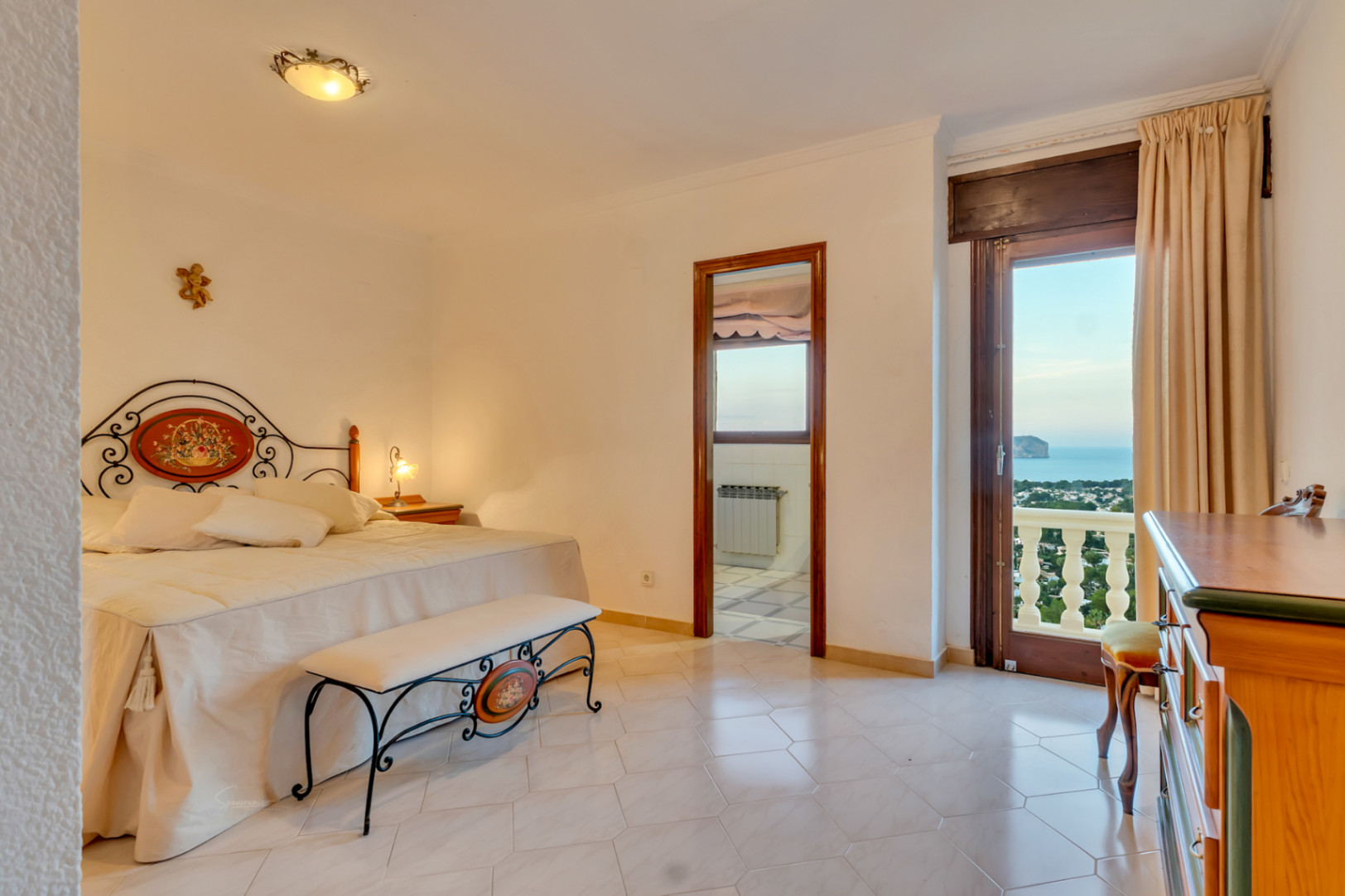 Slaapkamer met decoratief smeedijzeren hoofdbord, tegelvloer en toegang tot het balkon met panoramisch uitzicht op de baai en de Middellandse Zee.