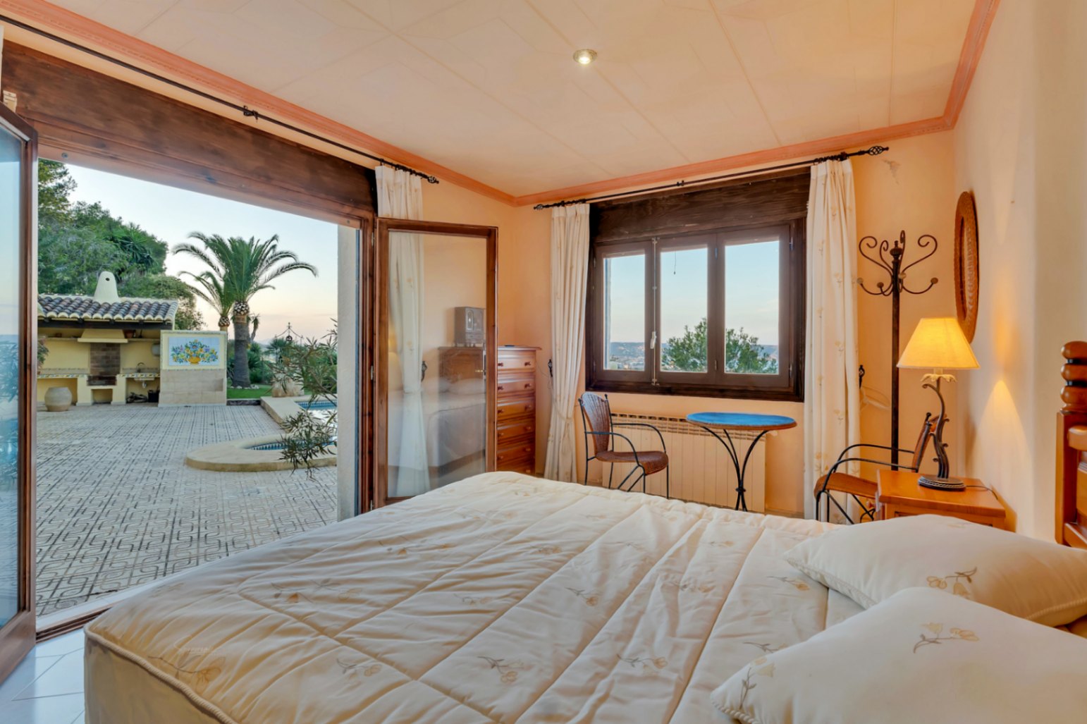 Slaapkamer met directe toegang tot het terras en zwembad via glazen deuren, donkerhouten kozijnen en uitzicht op de mediterrane tuin en barbecueplaats.