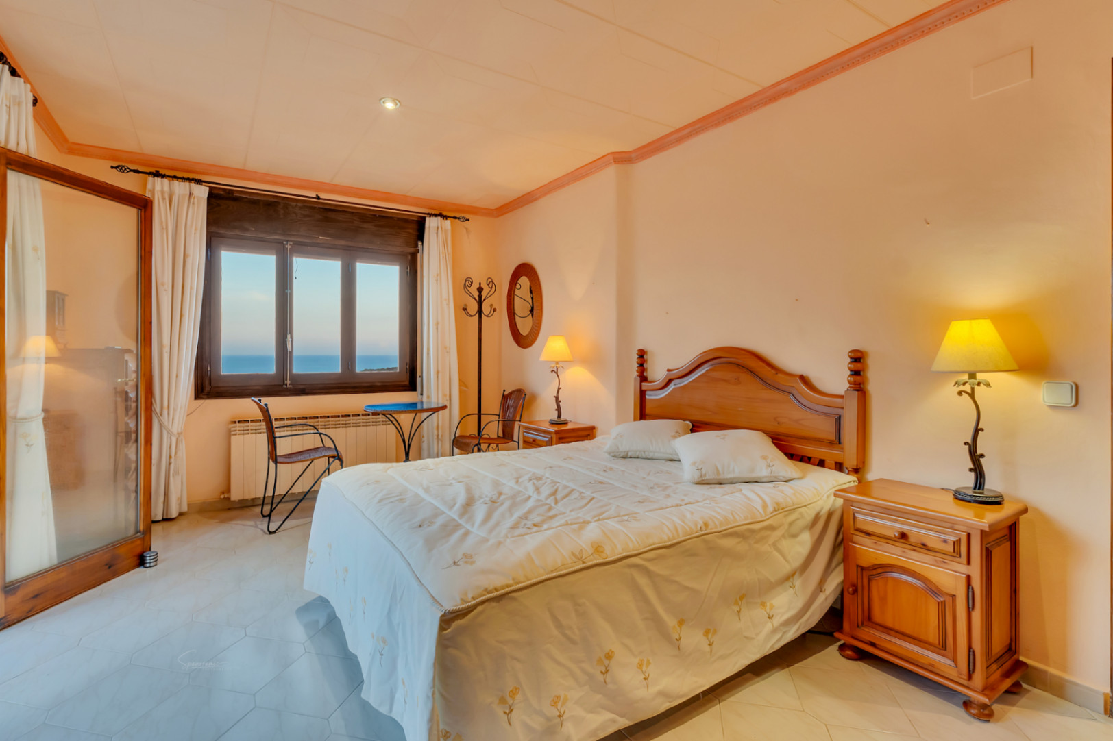 Slaapkamer met klassiek houten meubilair, zeshoekige vloertegels en een raam met uitzicht op zee. Voorzien van een radiator en een kleine zithoek bij het venster.