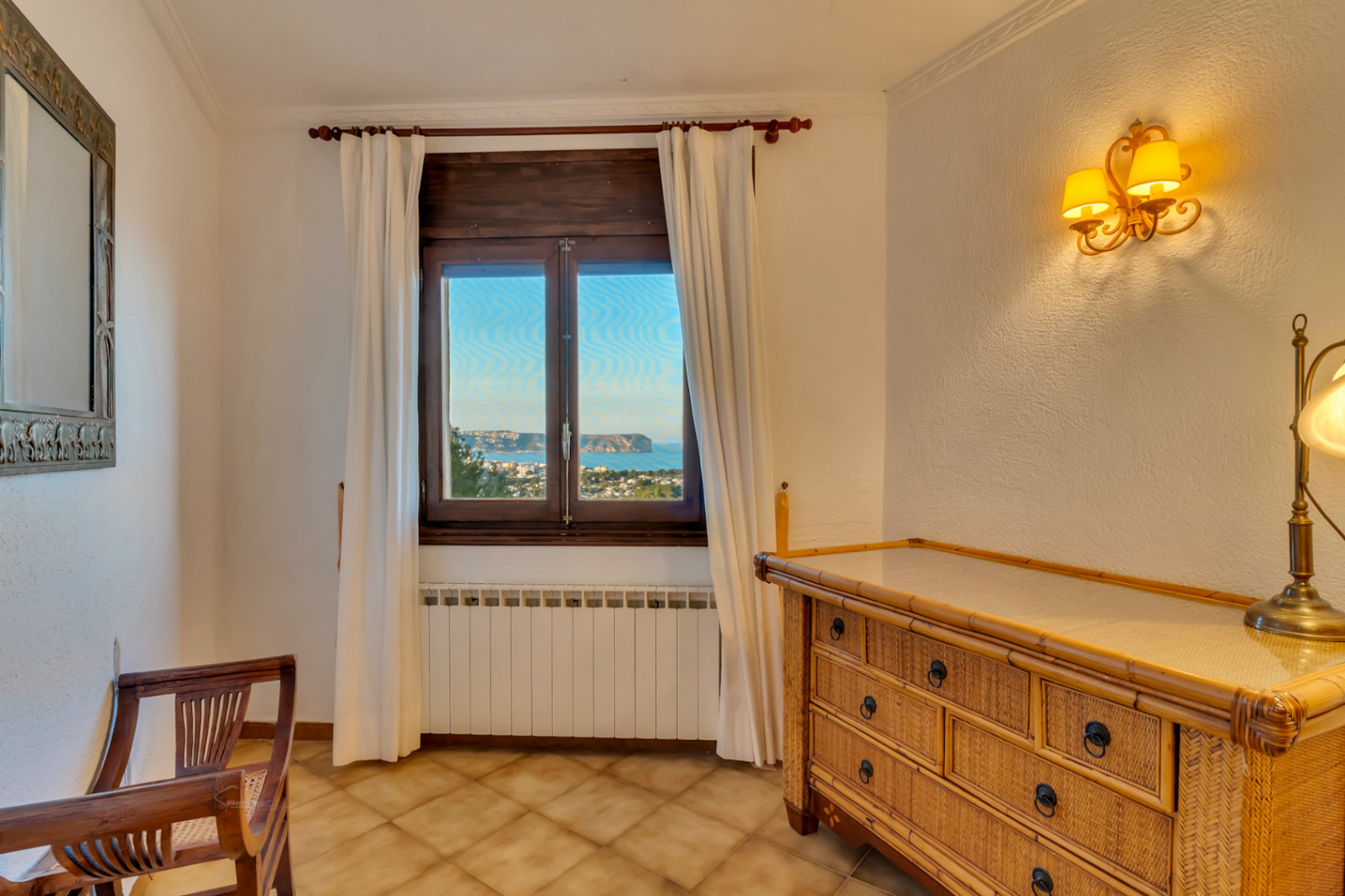 Slaapkamerhoek met rieten ladekast, houten stoel en raam met uitzicht op zee en Cabo San Antonio. Voorzien van radiator en sfeervolle wandverlichting.