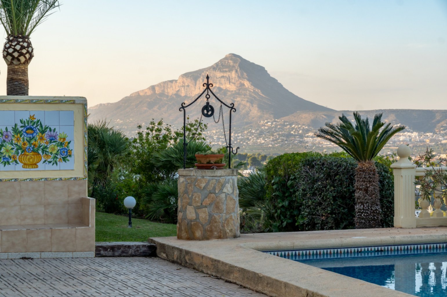 Terras bij het zwembad met een decoratieve stenen put en keramisch tableau, met panoramisch uitzicht op de Montgó en de kust van Jávea.