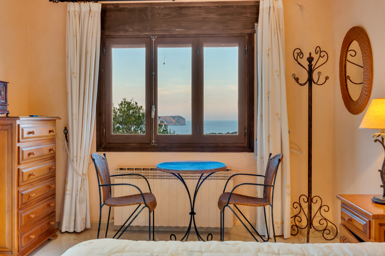 Zithoek in de slaapkamer met een houten raam dat uitkijkt op Cabo San Antonio. Inclusief houten ladekast, radiator en een rieten spiegel.