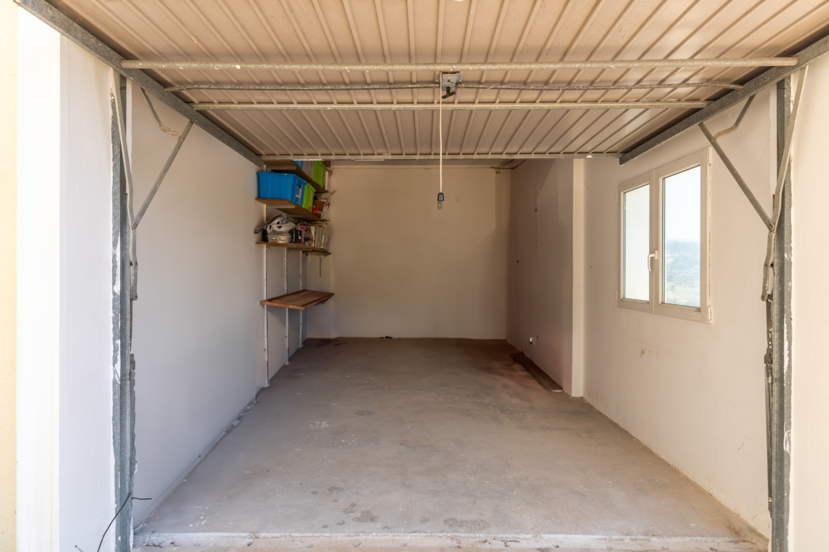 Afgesloten garage met betonvloer, witte muren en zijraam voor natuurlijk licht. Voorzien van opbergschappen aan de wand.