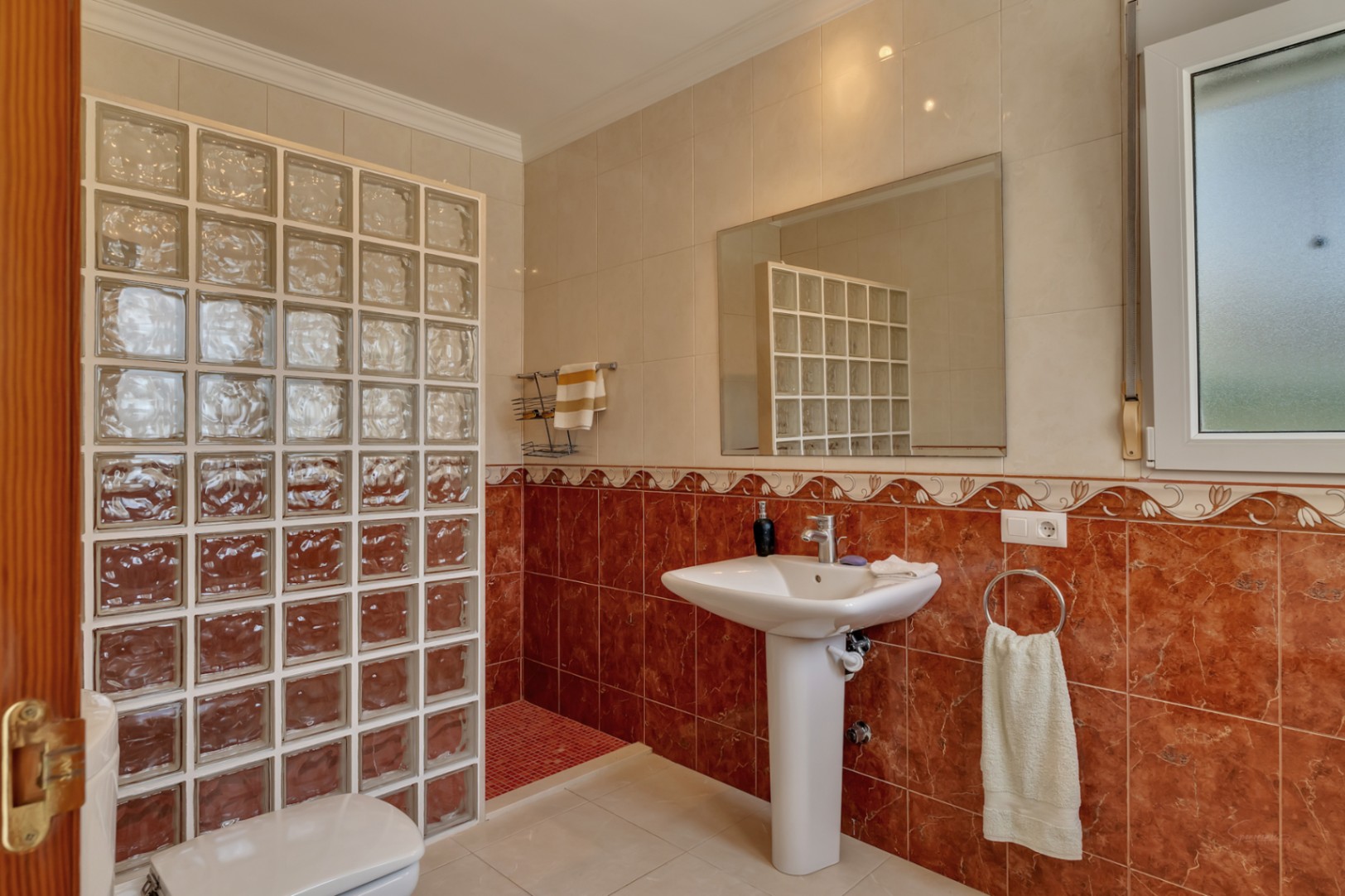 Badkamer met een kolomwastafel, grote spiegel en een inloopdouche gescheiden door een wand van glasbouwstenen en rode marmerlook tegels.