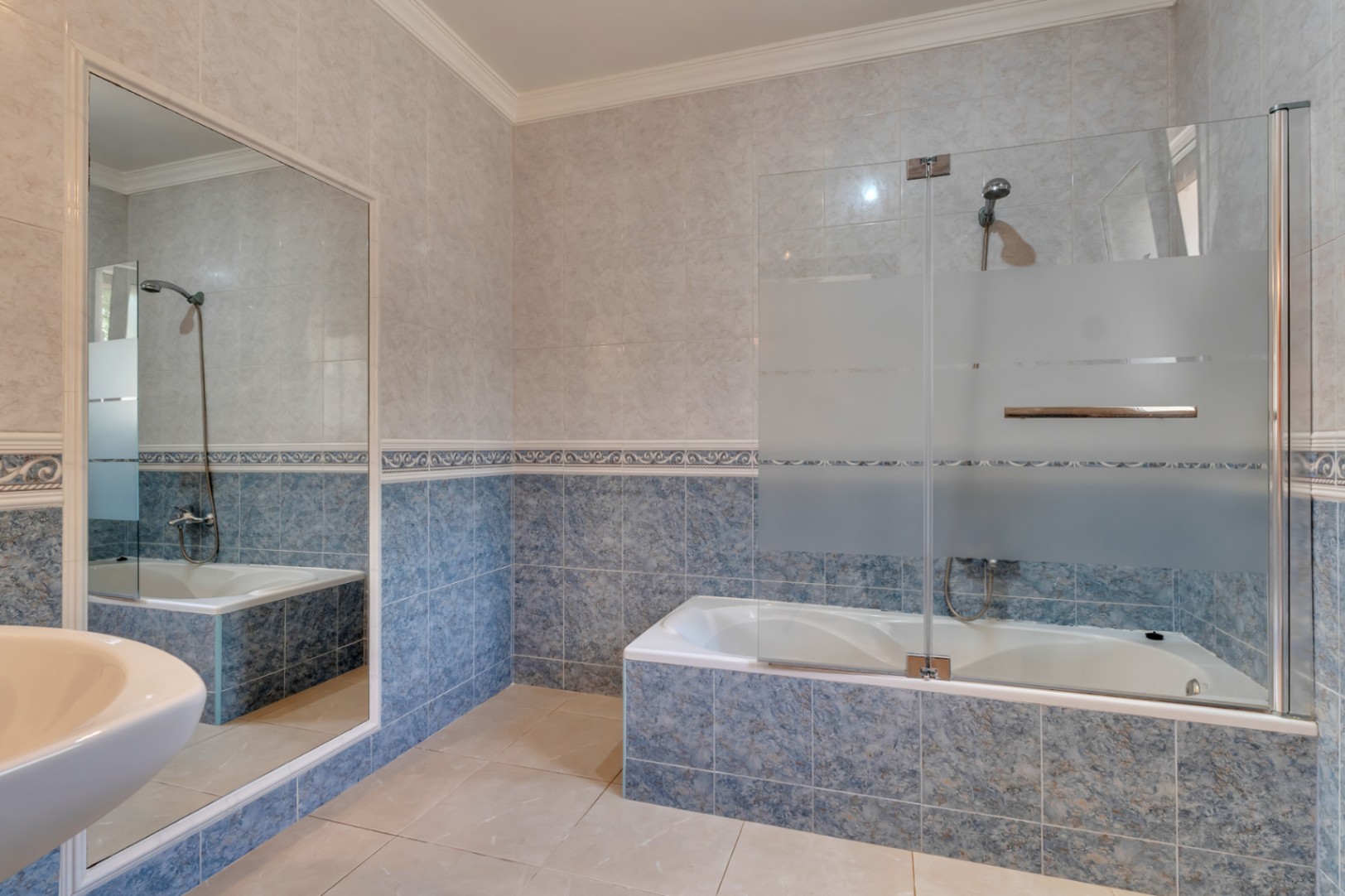 Badkamer met ingebouwd ligbad, glazen douchescherm, blauwe wandtegels met decoratieve sierrand en een grote wandspiegel.