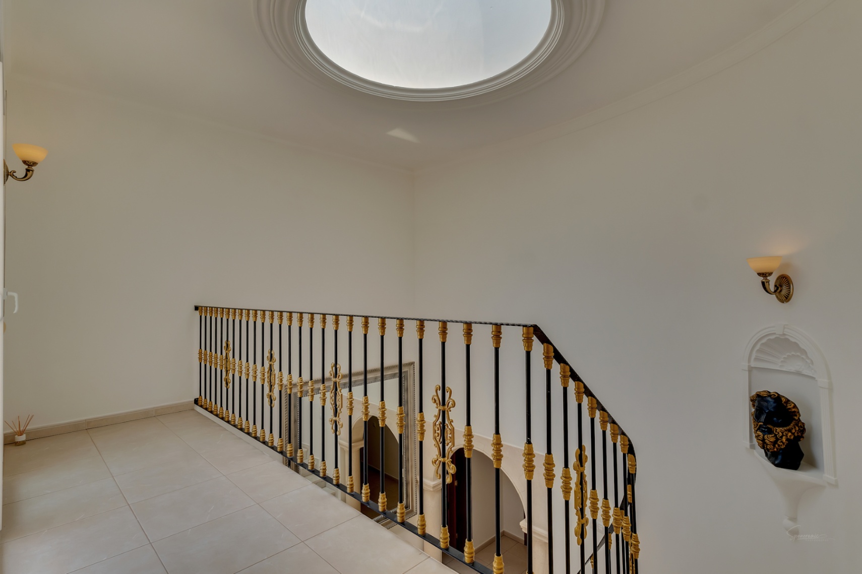 Bovenverdieping met ronde lichtkoepel, smeedijzeren balustrade met gouden accenten en een decoratieve muurnis. Zicht op de dubbelhoge hal.