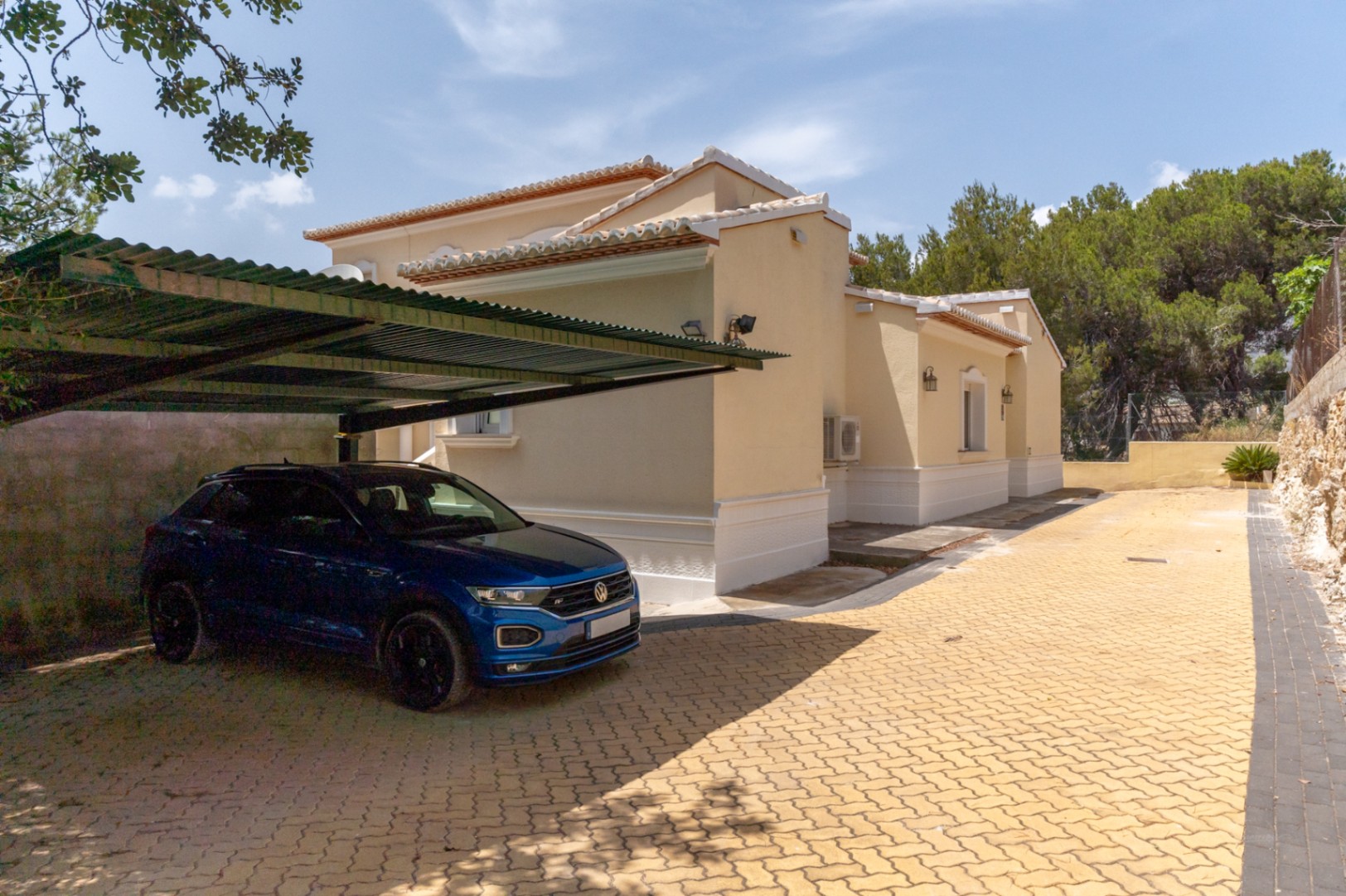 Buitenparkeerplaats met metalen carport en bestrating, gelegen naast de beige gevel van de villa met witte decoratieve elementen.