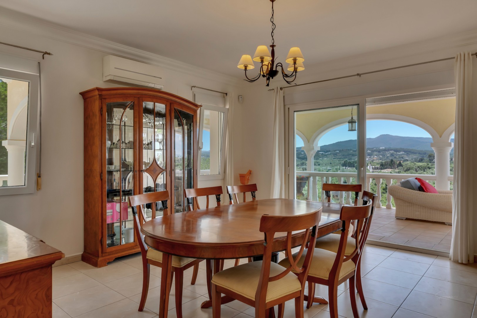 Eetkamer met een ovale houten tafel, klassieke vitrinekast en schuifpui naar het overdekte terras met panoramisch uitzicht over de vallei.