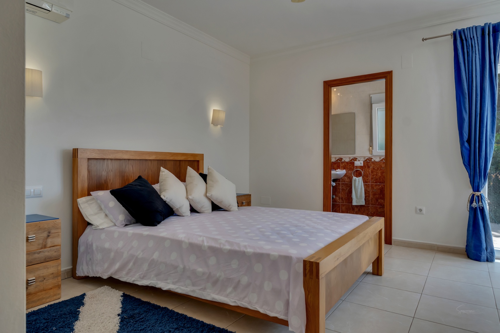 Lichte slaapkamer met massief houten bedframe, tegelvloer, airconditioning en een aangrenzende badkamer met roodachtige marmeren wandtegels.