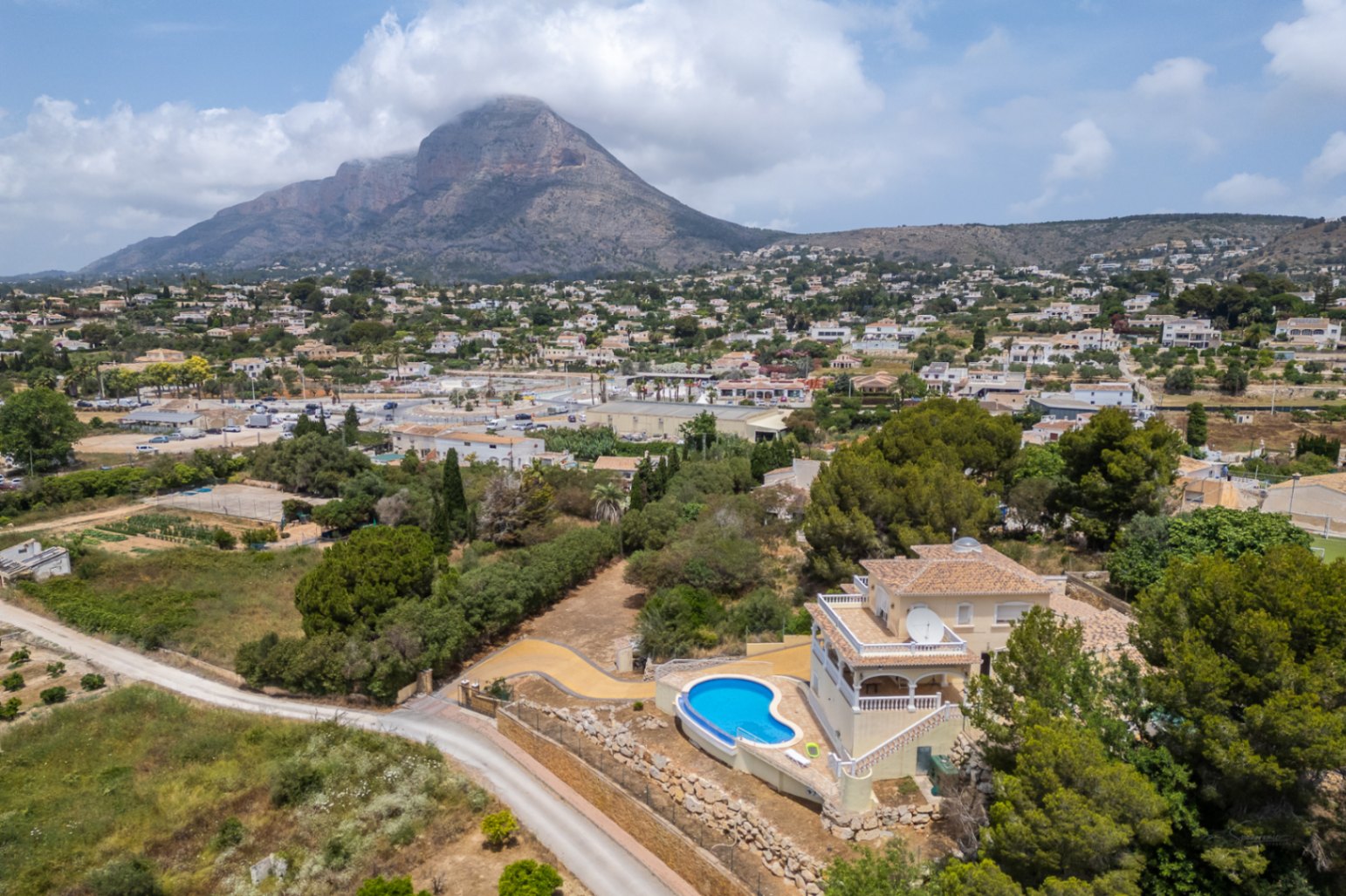 Luchtfoto van de villa met een gebogen overloopzwembad en verhoogde terrassen, uitkijkend over de vallei van Jávea en de berg Montgó.