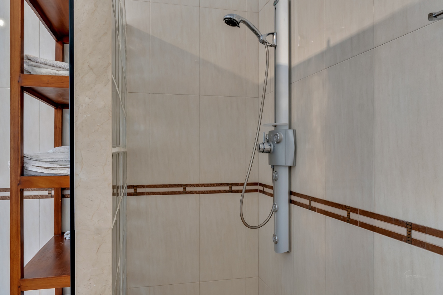 Moderne douche met hydromassagekolom, crème keramische tegels met decoratieve rand en houten opbergrek naast een wand van glasbouwstenen.