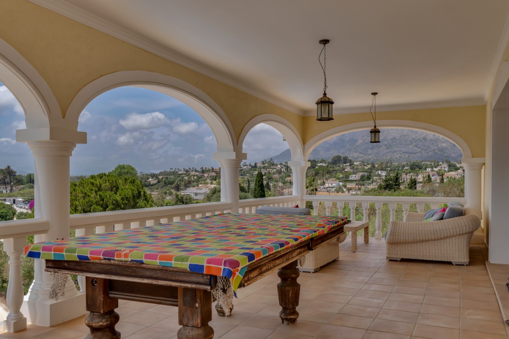 Overdekt terras met klassieke bogen en witte kolommen, voorzien van een biljarttafel en zithoek, met panoramisch uitzicht op de Montgó en de vallei.