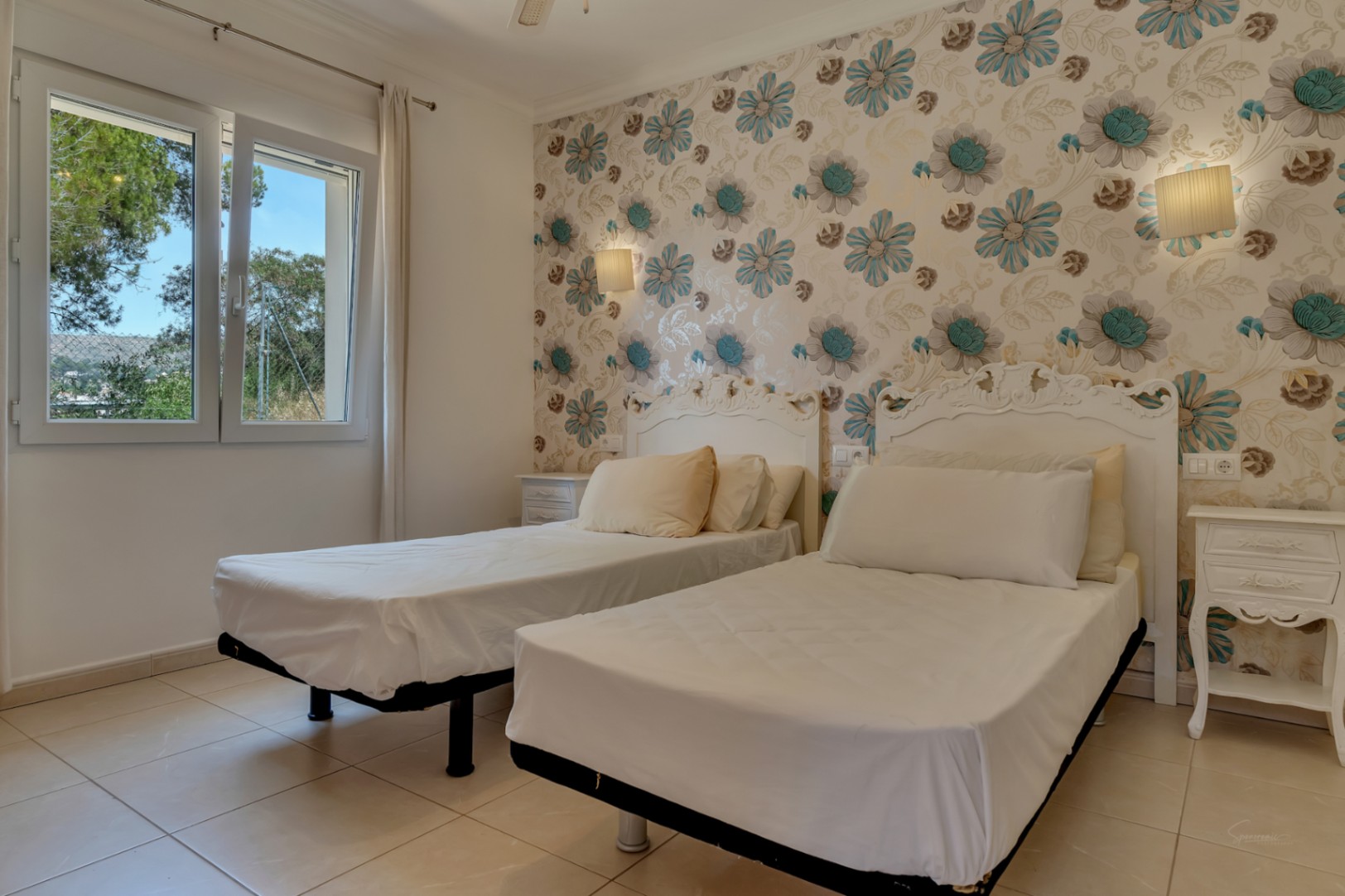 Slaapkamer met twee eenpersoonsbedden, sierlijke witte hoofdeinden en bloemenbehang. Voorzien van een tegelvloer en een raam met uitzicht op de groene omgeving.