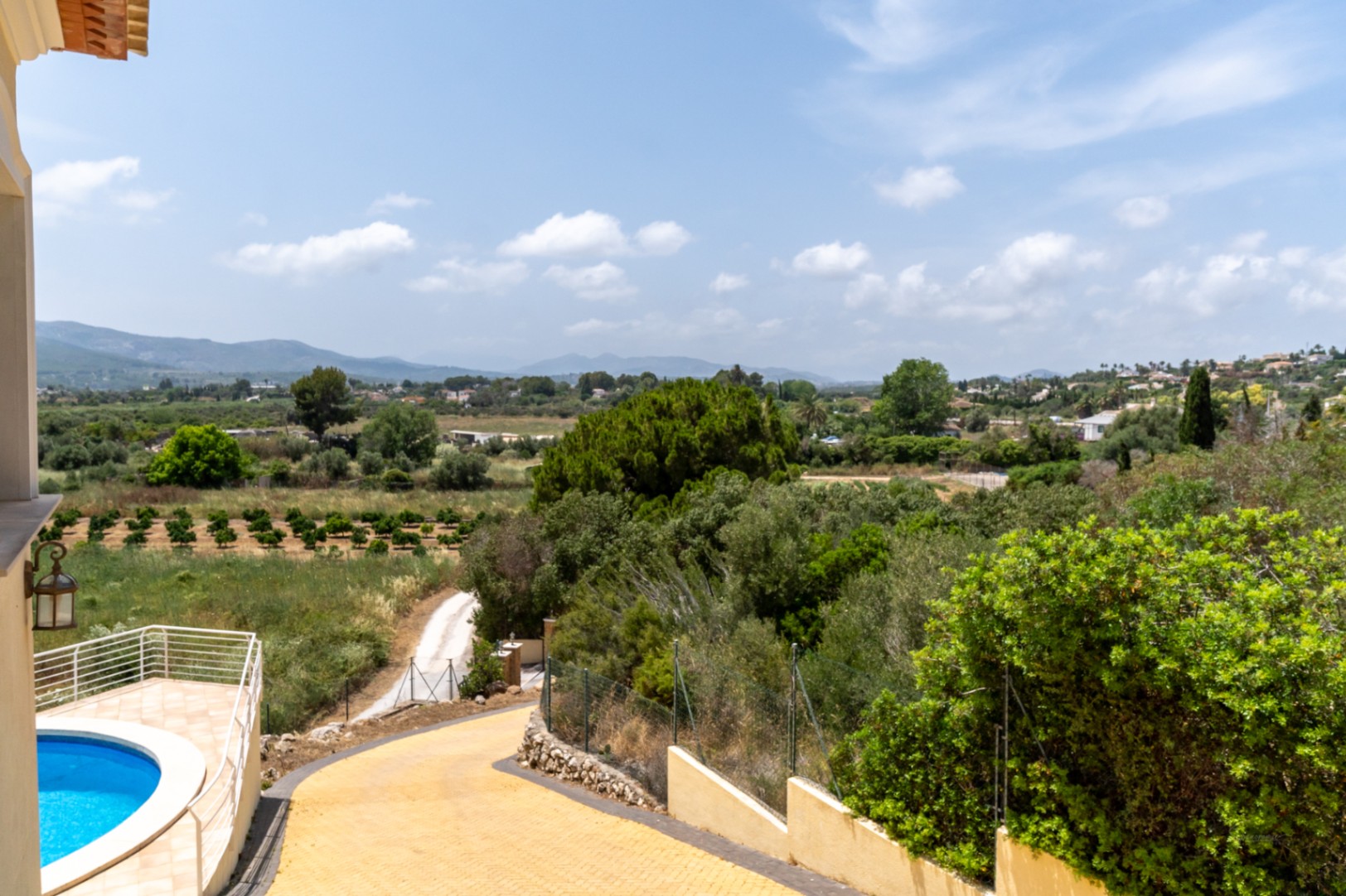 Verhoogd uitzicht op de oprit, het ronde zwembad met wit hekwerk en het panoramische zicht over de vallei van Jávea met uitgestrekte sinaasappelgaarden.