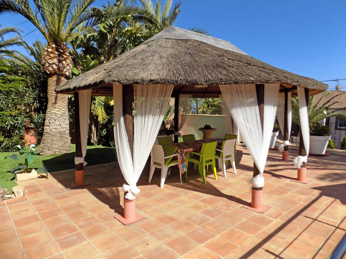 Buitenprieel met rieten dak en witte gordijnen op een terracotta terras, gelegen in een mediterrane tuin met palmbomen en een eethoek.