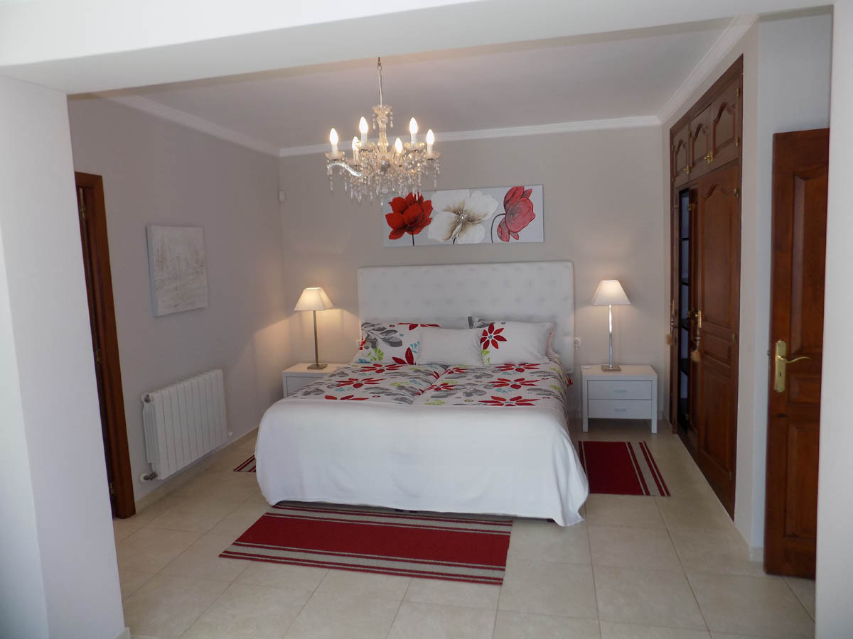 Slaapkamer met houten inbouwkasten, wit gecapitonneerd hoofdbord, kristallen kroonluchter en lichte tegelvloer met radiator.