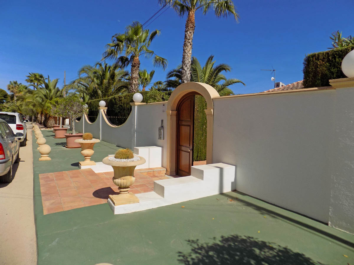Voetgangersingang met een houten boogdeur, witte omheiningsmuur gedecoreerd met stenen vazen en cactussen, omgeven door palmbomen en mediterrane beplanting.