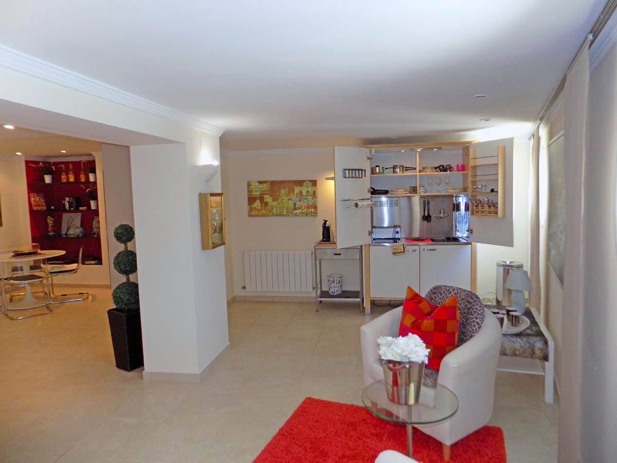 Woonkamer op de begane grond met een compacte open kitchenette, zithoek met rood tapijt en lichte tegelvloer.