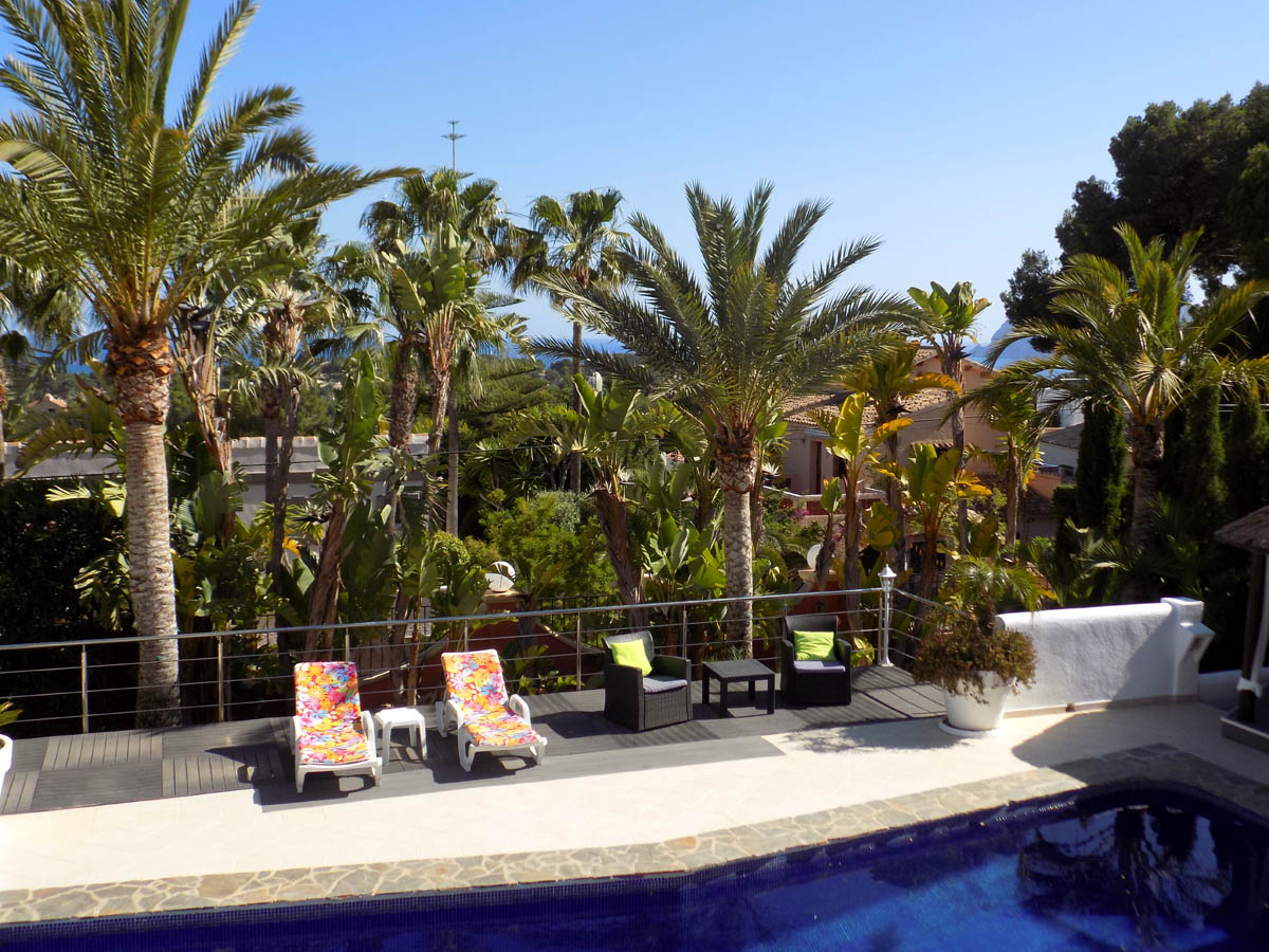 Zonneterras met houten vlonder en metalen balustrade bij het zwembad, omgeven door volgroeide palmbomen en mediterrane beplanting met uitzicht op zee.