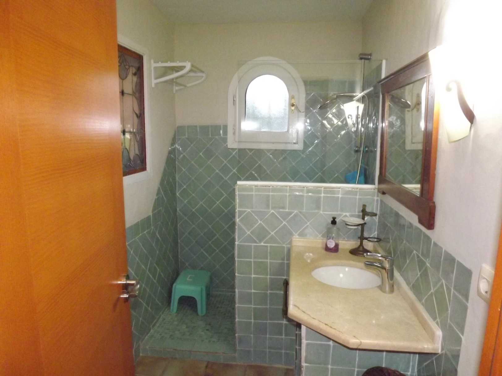Badkamer met groene tegels, natuurstenen wastafelblad en inloopdouche met boogvenster en glazen wand. Voorzien van een decoratief glas-in-loodraam.