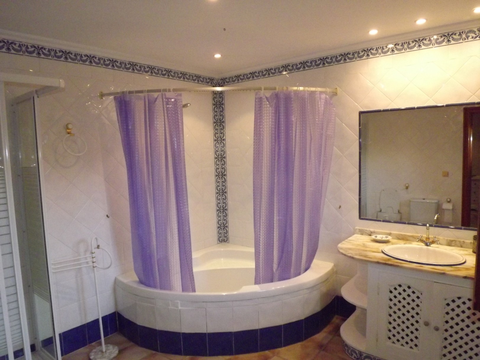Badkamer met hoekbad, paarse gordijnen en witte tegels met blauwe sierranden. Voorzien van een wastafelmeubel met marmeren blad en grote spiegel.
