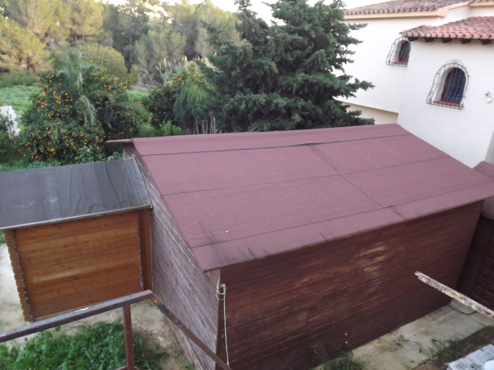 Houten berging met vilten dak op het grote perceel, omringd door mediterrane beplanting en sinaasappelbomen nabij de hoofdwoning.
