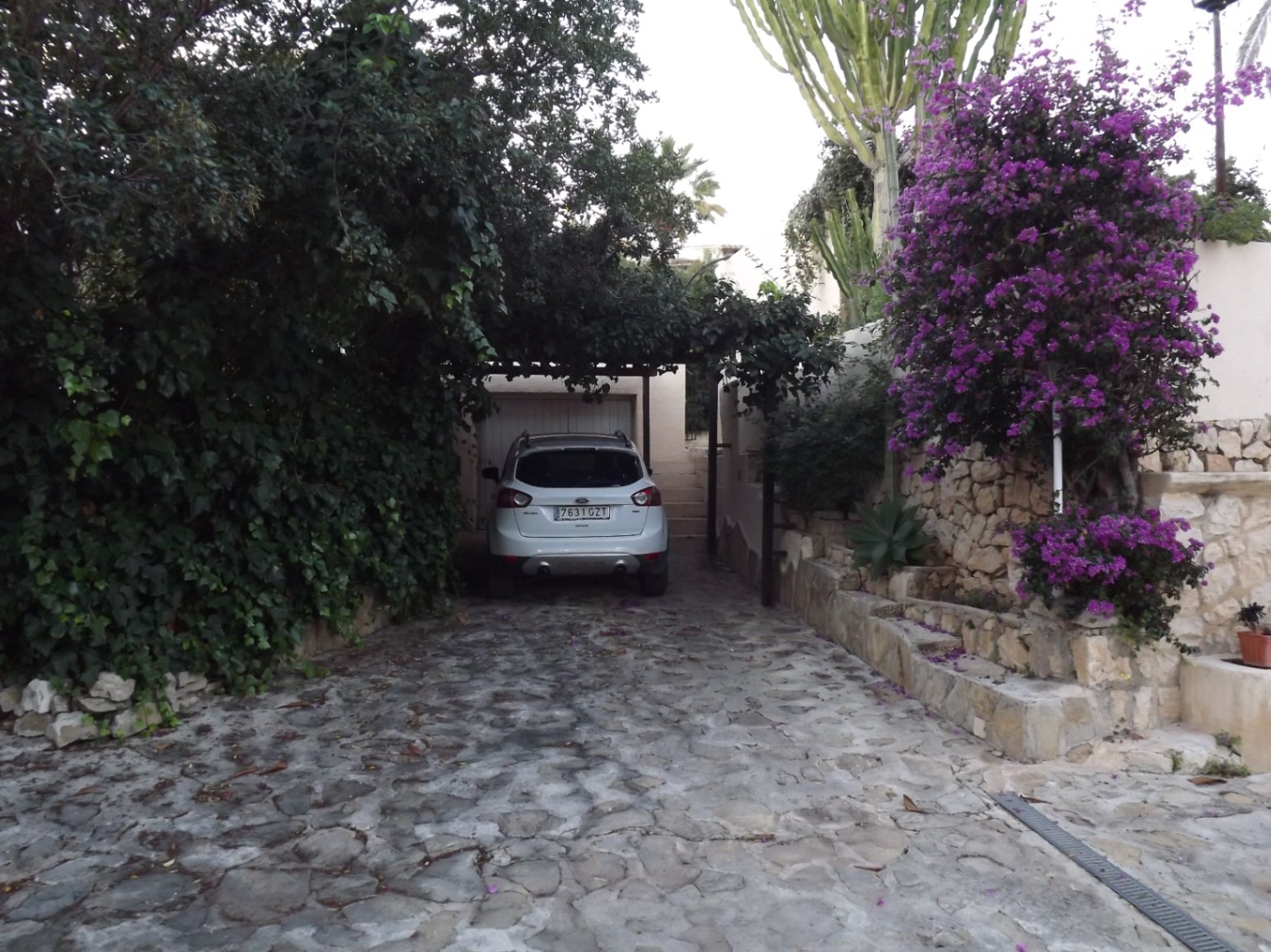 Oprit van natuursteen naar de garage, omgeven door weelderig mediterraan groen, paarse bougainville en traditionele droge stenen muren.