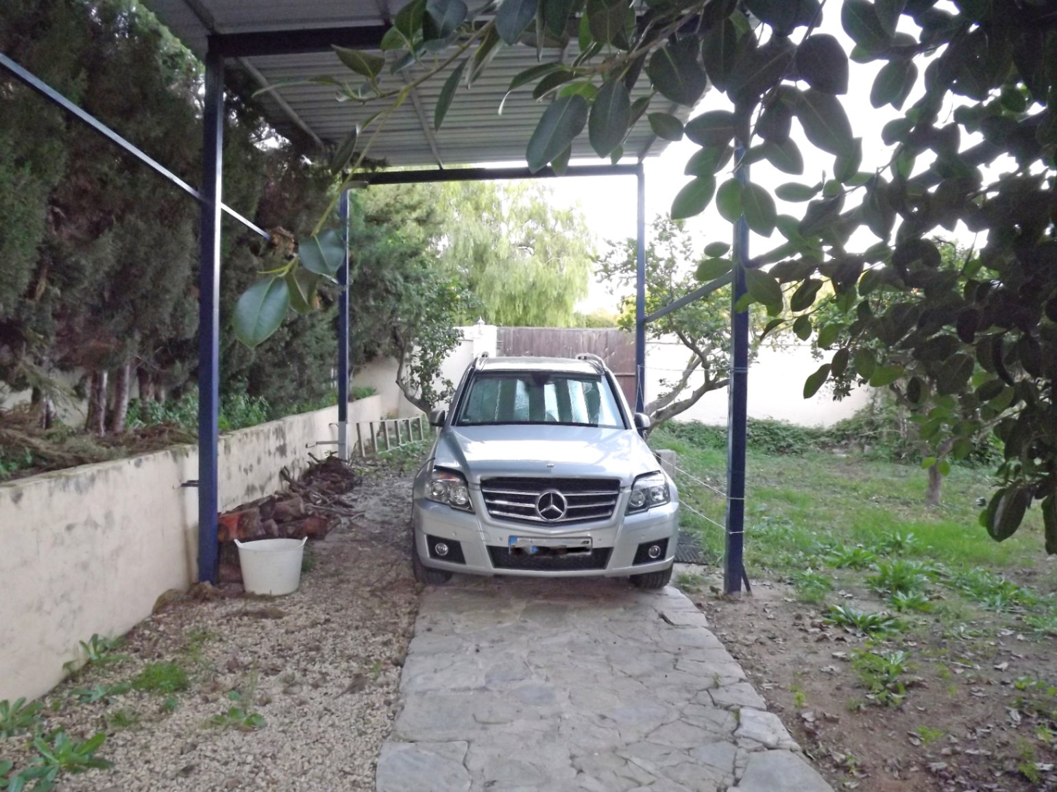 Overdekte parkeerplaats voorzien van een metalen carport en een oprit van natuursteen, omgeven door volwassen bomen en grind.