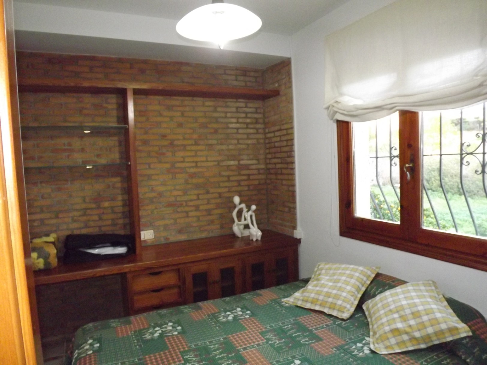 Slaapkamer met een accentmuur van schoon metselwerk, ingebouwde houten kasten met glazen planken en een houten raamkozijn met uitzicht op de tuin.