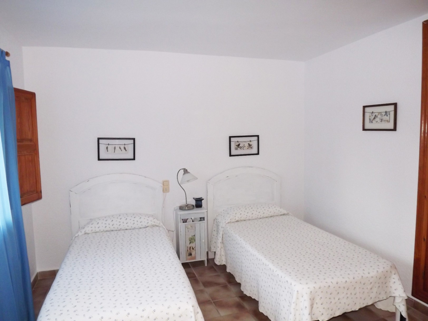 Slaapkamer met twee eenpersoonsbedden, witte hoofdborden en terracotta tegelvloer. Voorzien van een nachtkastje en een raam met houten luiken.