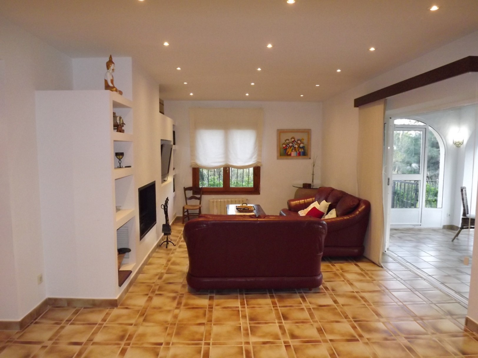 Woonkamer met bordeauxrode lederen banken, een ingebouwde open haard en inbouwspots. De rustieke tegelvloer biedt toegang tot het glazen terras.
