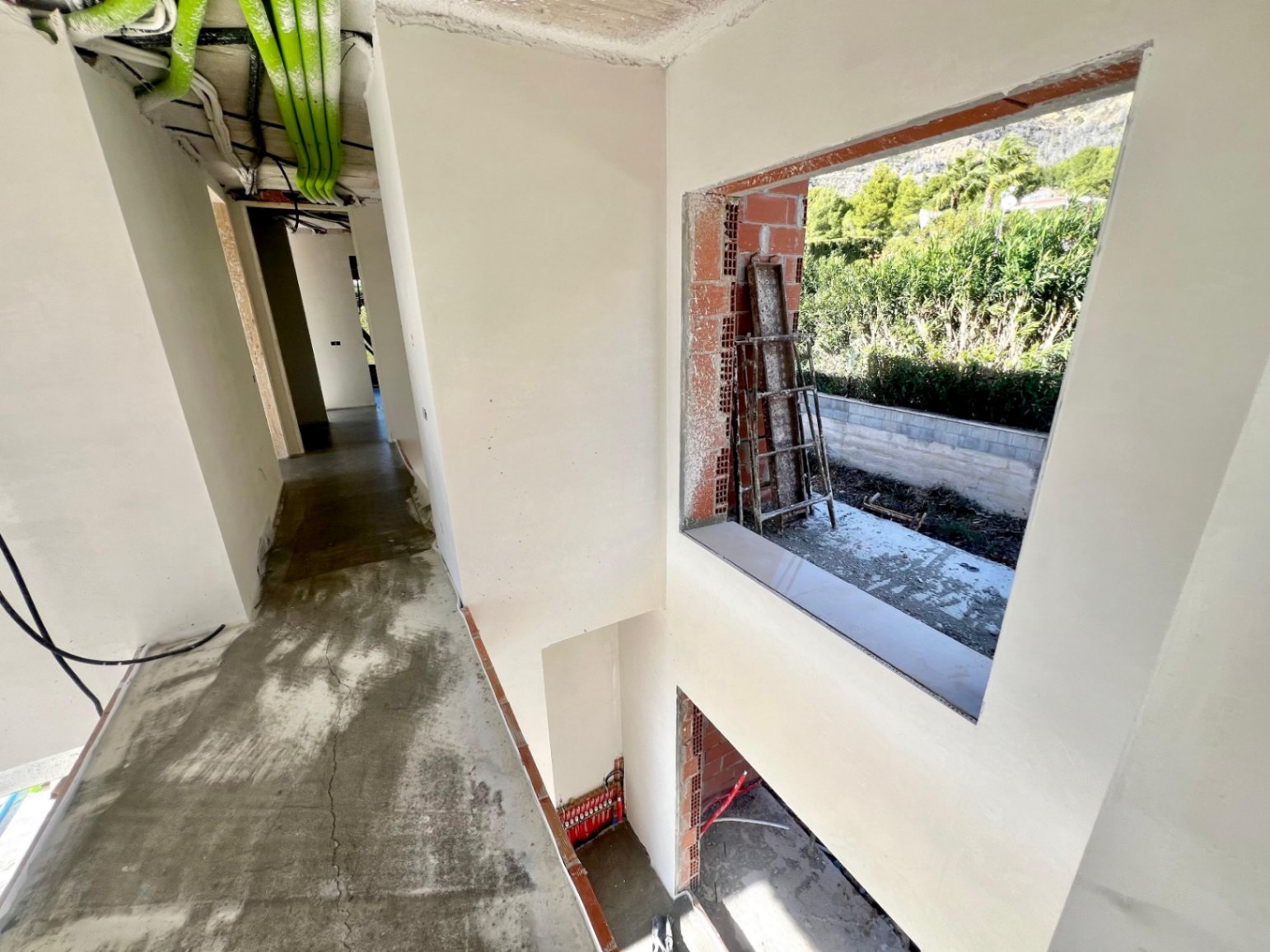 Binnengang in de bouwfase met dubbelhoge plafonds, zichtbare technische installaties en een grote raamopening naar de natuurlijke omgeving.