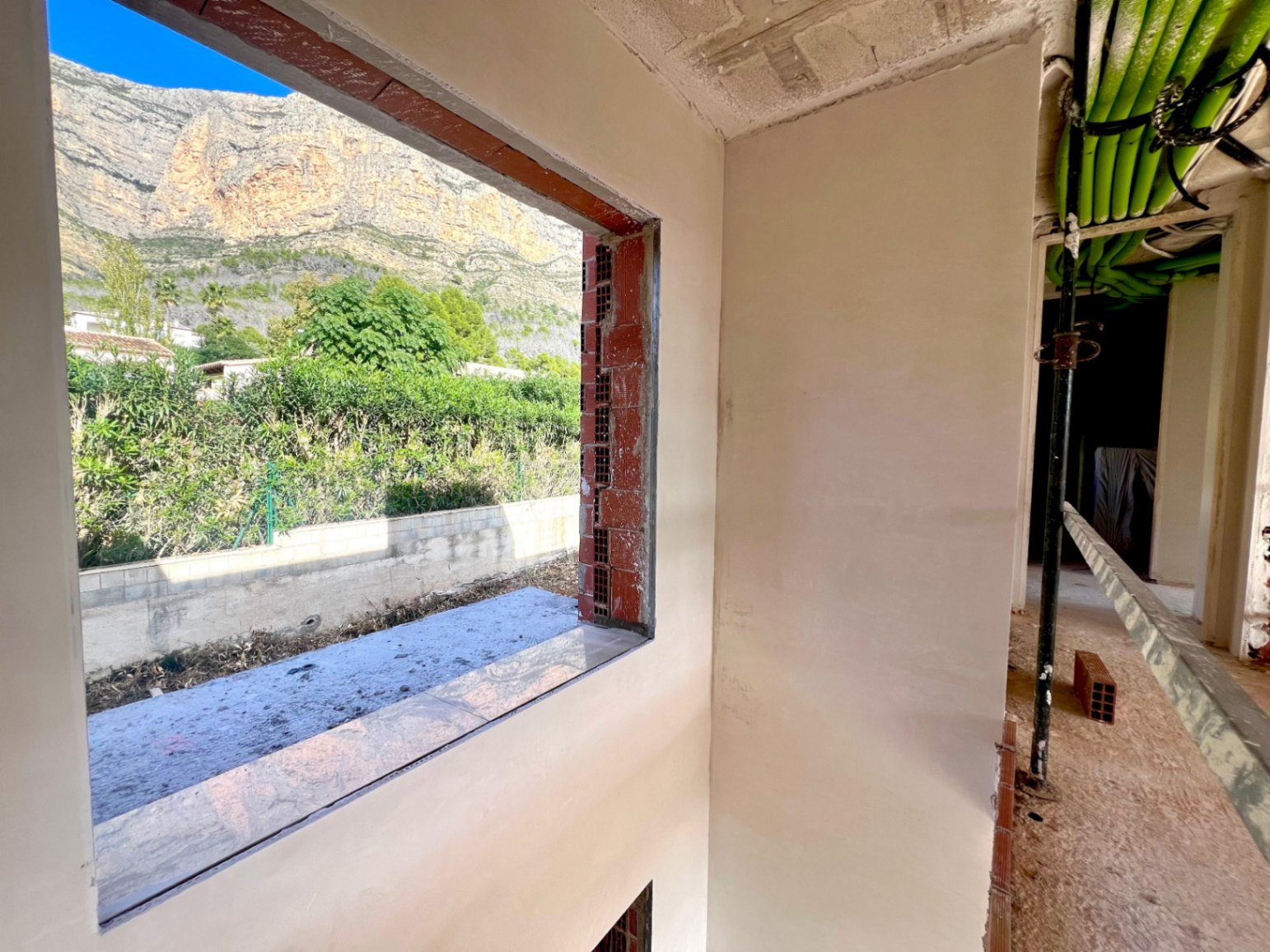 Interieur in aanbouw met een groot raamkozijn dat uitkijkt op de Montgó-berg, inclusief zichtbare technische leidingen aan het plafond en gestuukte muren.