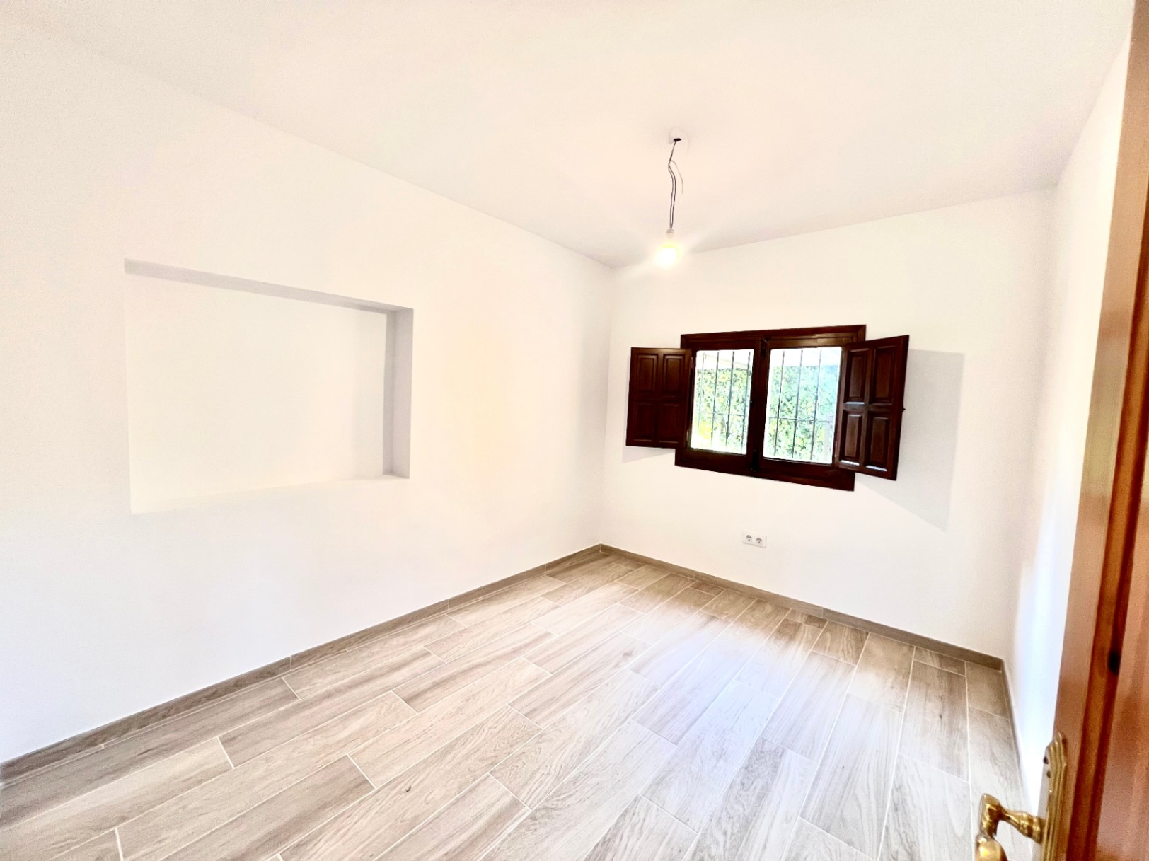 Lichte kamer met houtlook tegelvloer, een decoratieve muurnis en een donkerhouten raam met traditionele binnenluiken en tralies.