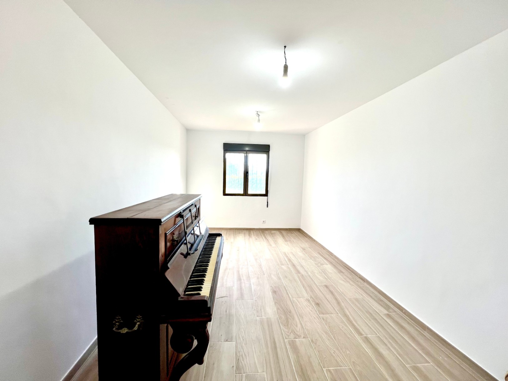 Lichte kamer met witte muren, keramische vloertegels met houtlook, een klassieke buffetpiano en een raam met een donker kozijn.