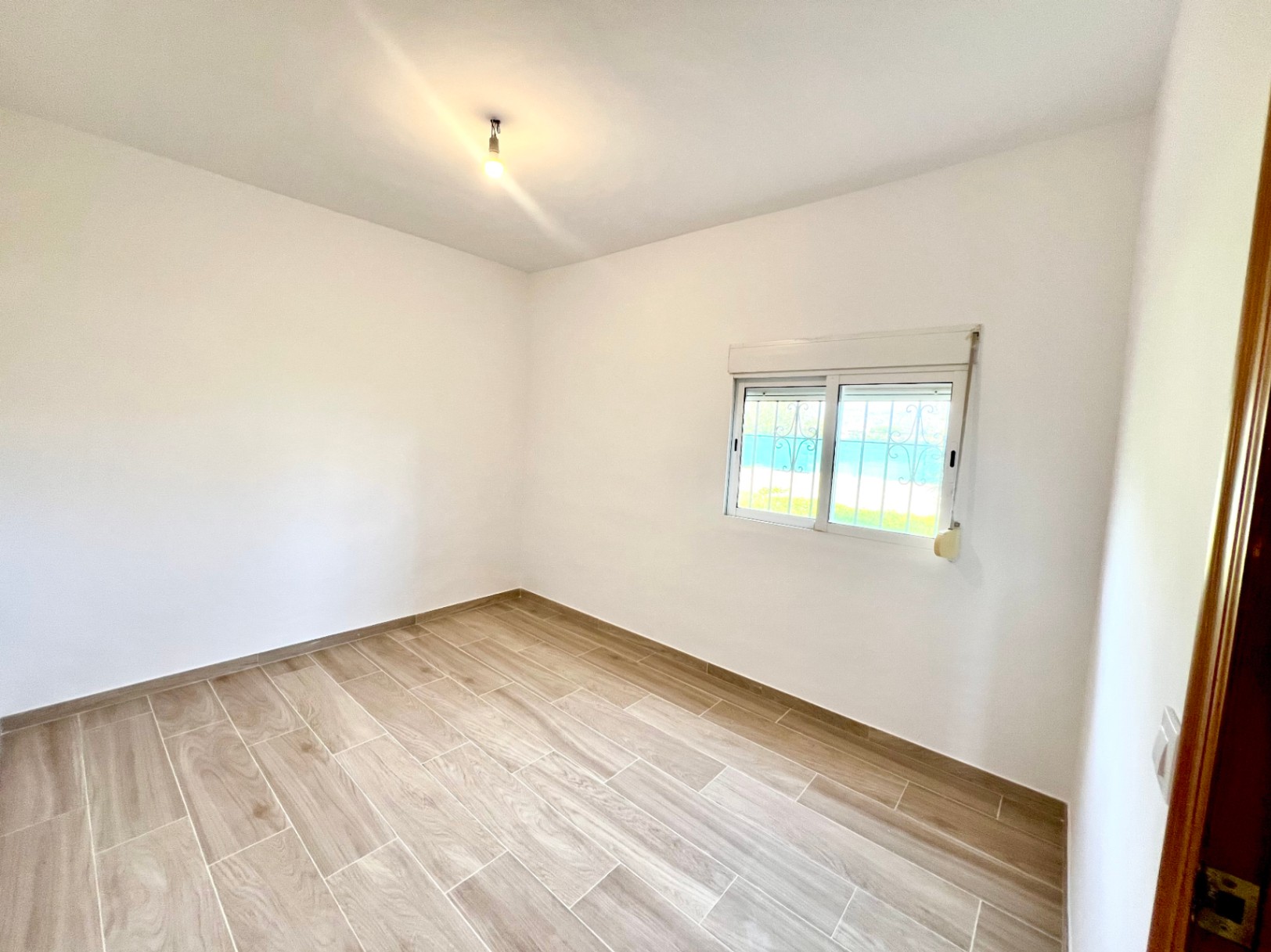 Lichte slaapkamer met houtlook keramische tegelvloer, strakke witte muren en een raam voorzien van sierlijk veiligheidshekwerk.