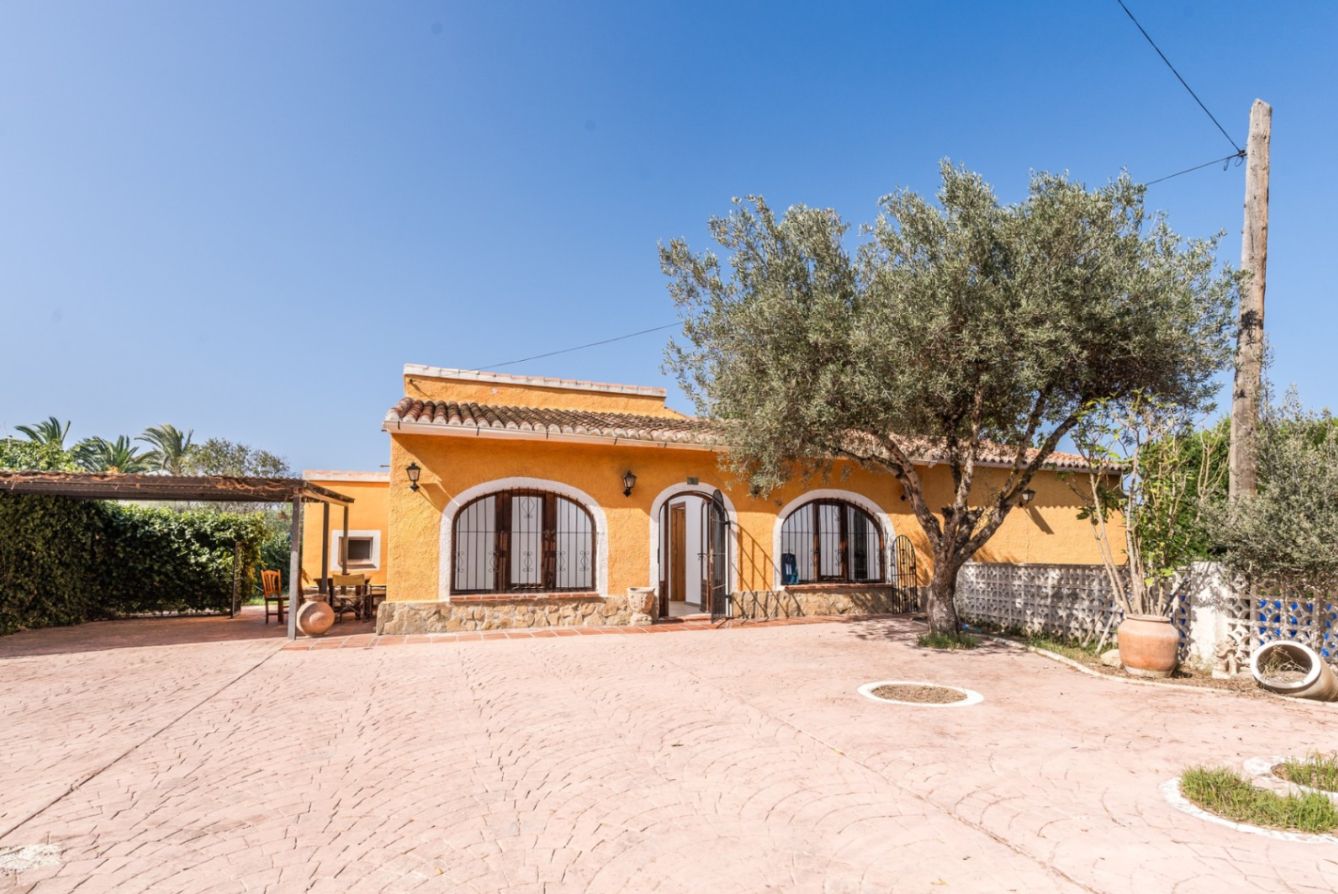 Mediterrane gevel met okerkleurige muren, boogvensters en een natuurstenen plint, grenzend aan een ruim bestraat terras met een volwassen olijfboom.