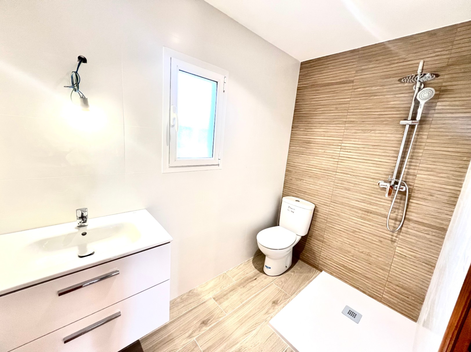 Moderne badkamer met inloopdouche, houtlook tegels, een wit wastafelmeubel en natuurlijke lichtinval via het zijraam.