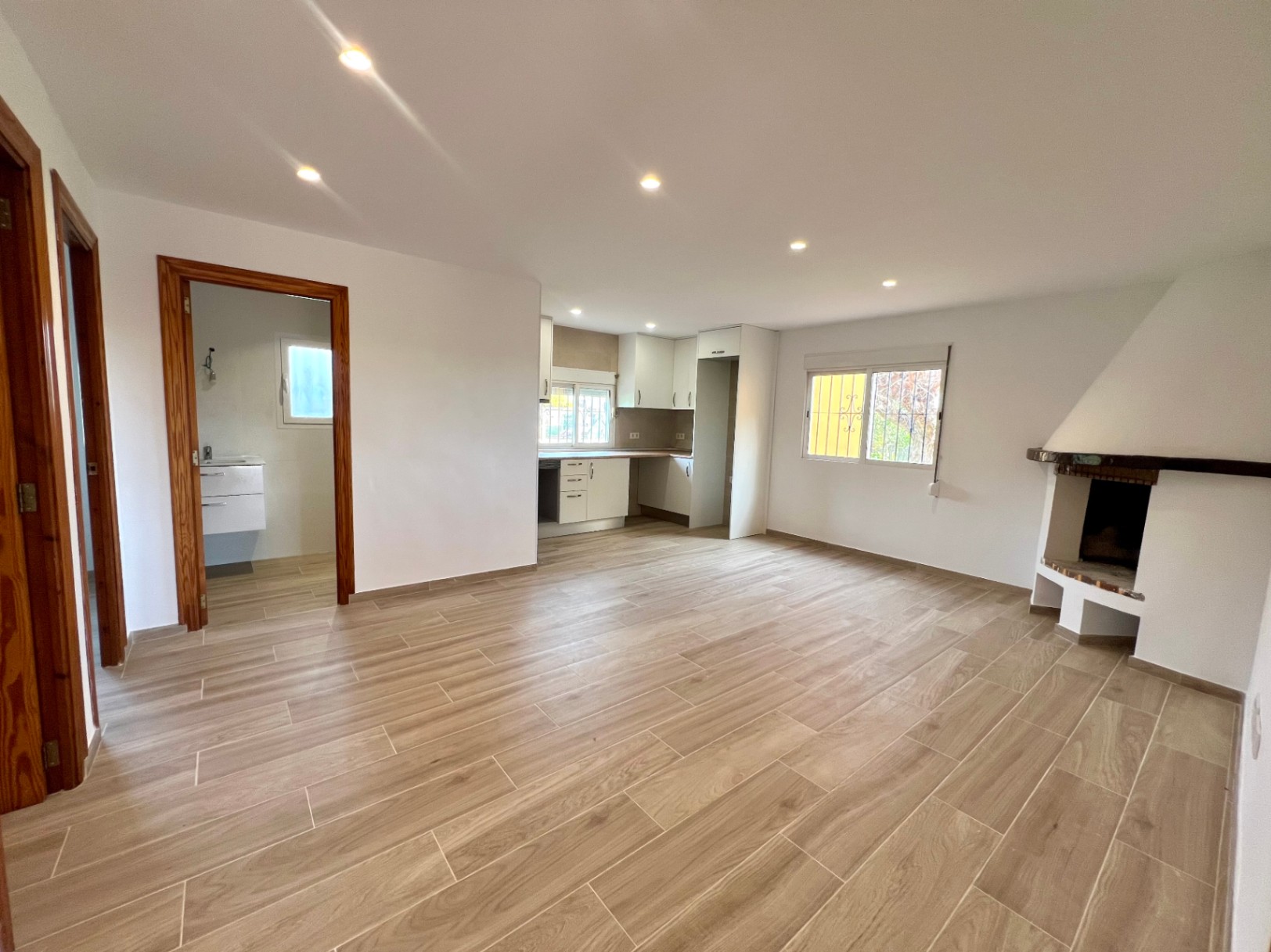 Open woonruimte met hoekhaard, houtlook tegelvloer en een witte inbouwkeuken. Voorzien van moderne inbouwspots en kozijnen van natuurlijk hout.