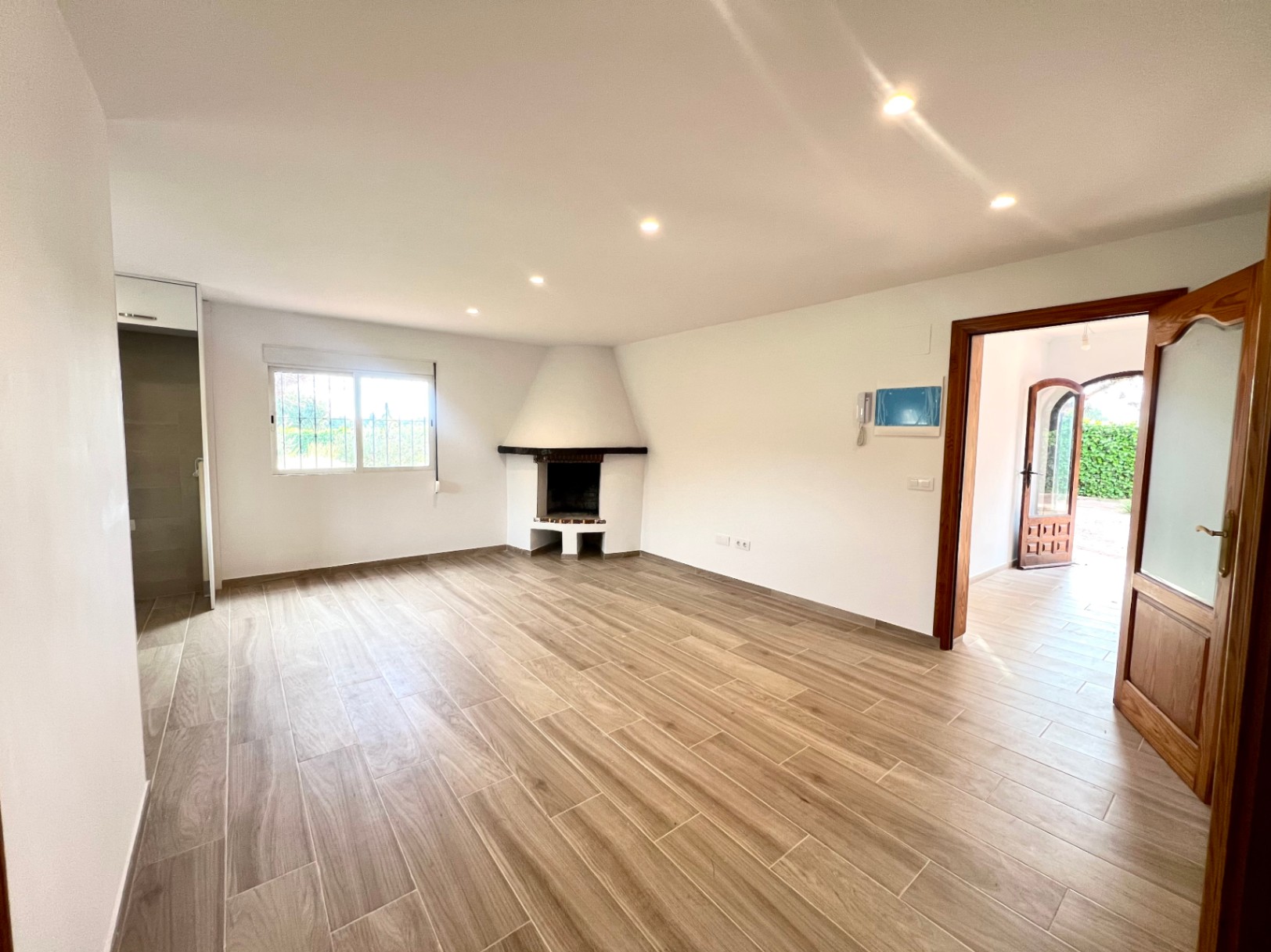 Ruime woonkamer met hoekhaard, keramische vloer met houtlook en inbouwspots. De ruimte staat in verbinding met de hal en biedt zicht op de tuin.